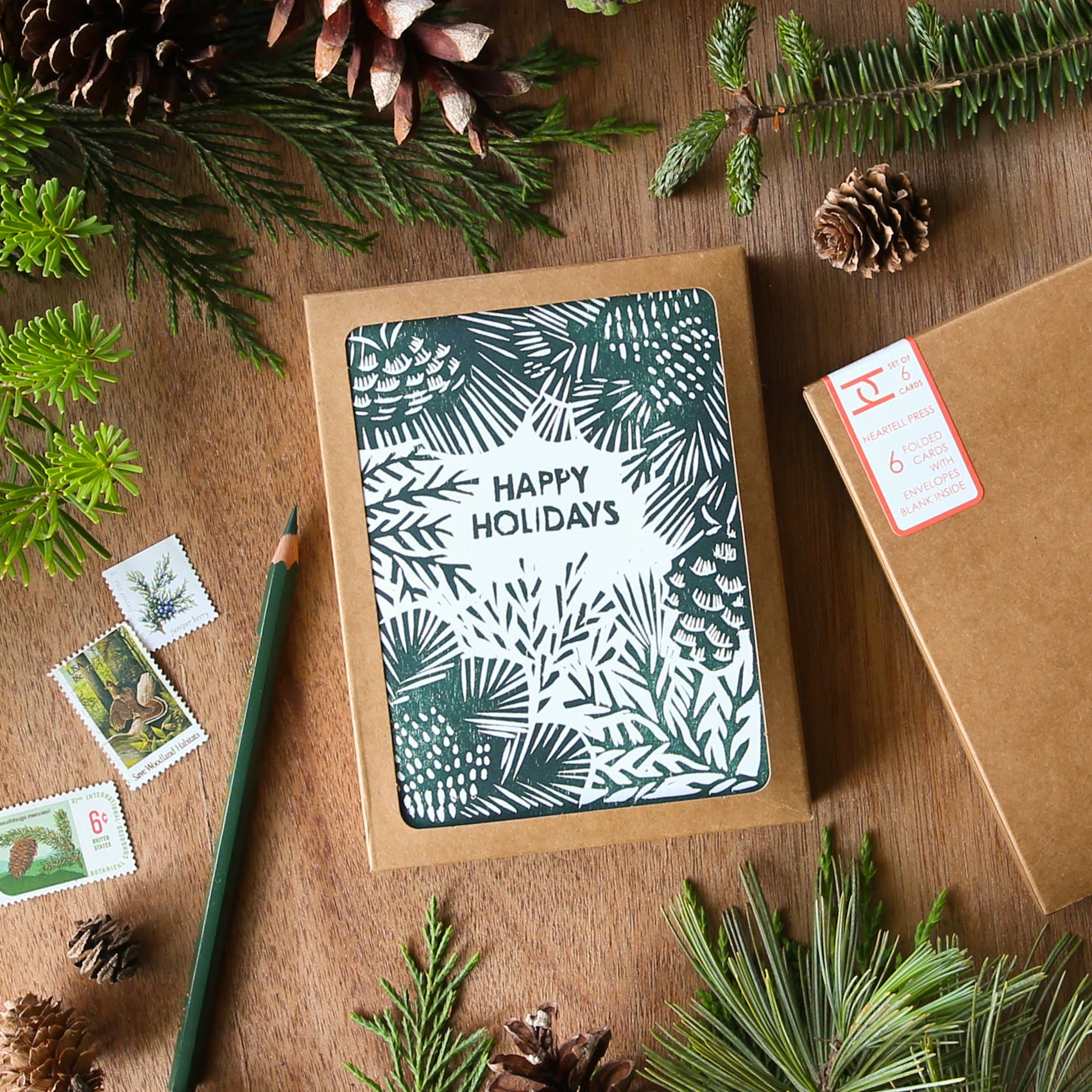 A square kraft paper gift box with a festive 'Happy Holidays' card insert featuring a dark green and white pinecone and leaf pattern, styled on a wooden tabletop with pine branches, pinecones, postage stamps, and a green pencil.