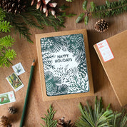 A square kraft paper gift box with a festive 'Happy Holidays' card insert featuring a dark green and white pinecone and leaf pattern, styled on a wooden tabletop with pine branches, pinecones, postage stamps, and a green pencil.