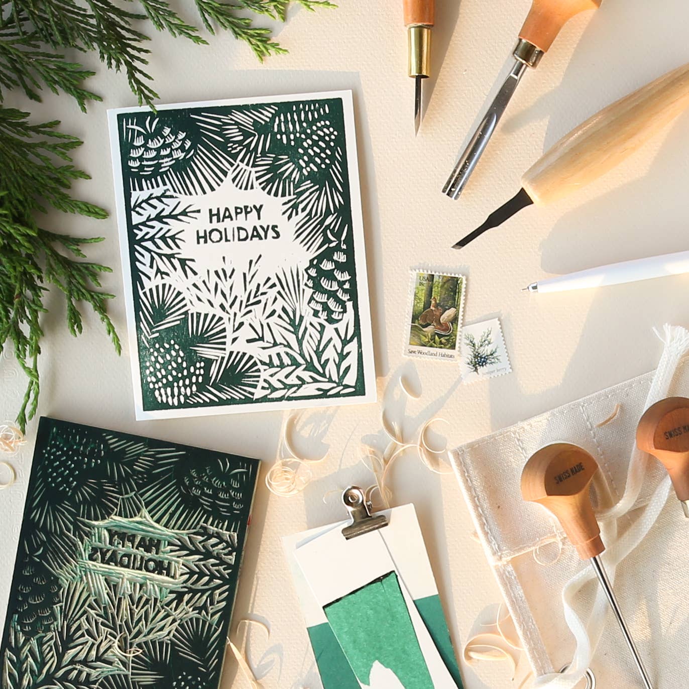 A styled flat-lay of handmade holiday greeting cards with dark green and white leafy pine designs, surrounded by carving tools, ribbon, and evergreen branches on a light beige surface.