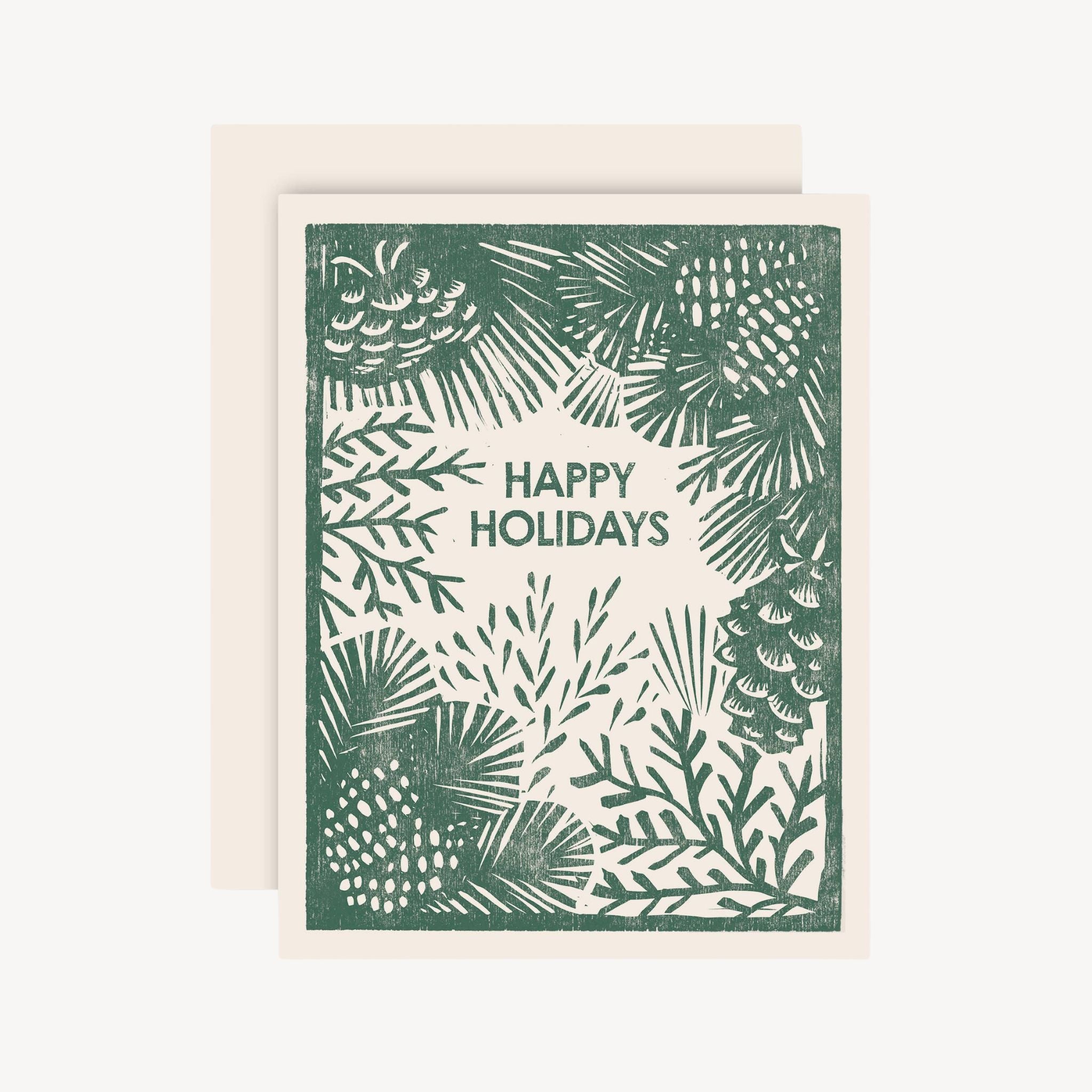 A holiday greeting card with a cream background and a dark green block-print style border of pine branches, pinecones, and the words “HAPPY HOLIDAYS” in the center. A matching cream envelope is partially visible behind the card on a white background.