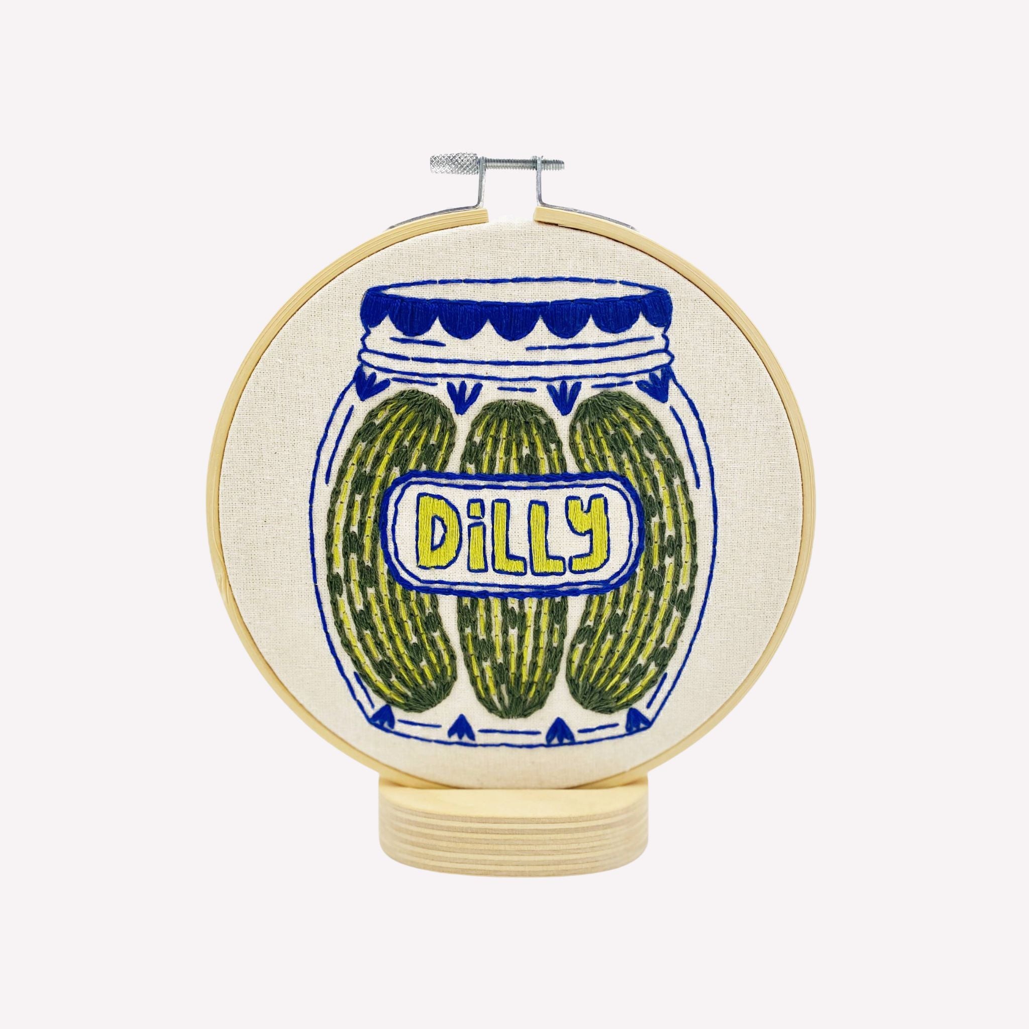 Embroidery hoop with a design of pickles in a jar labeled 'Dilly' on a white background