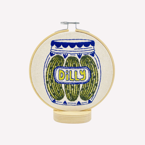 Embroidery hoop with a design of pickles in a jar labeled 'Dilly' on a white background