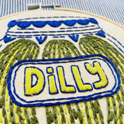 Embroidery hoop with a jar of pickles and the word 'Dilly' on a striped fabric background