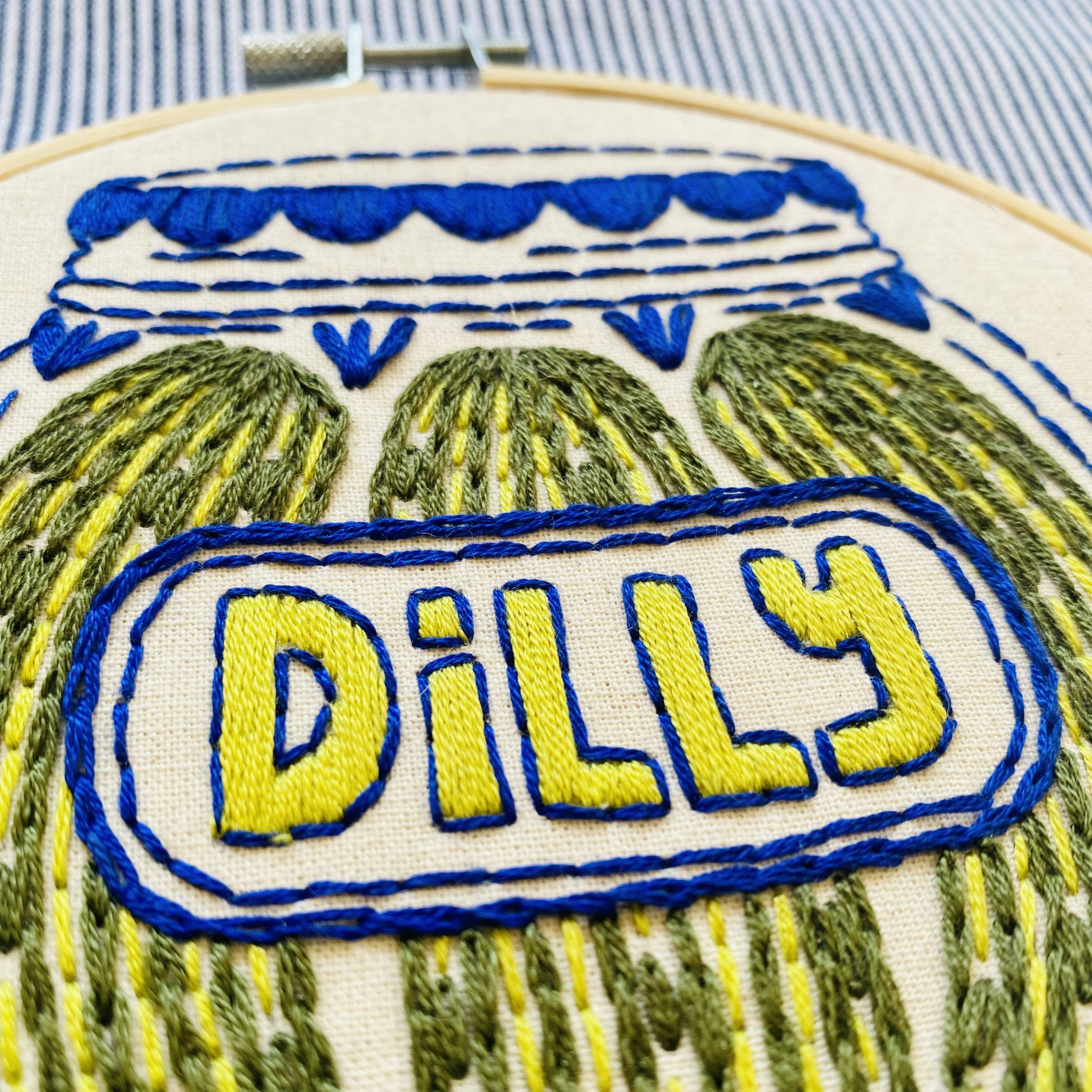 Embroidery hoop with a jar of pickles and the word 'Dilly' on a striped fabric background