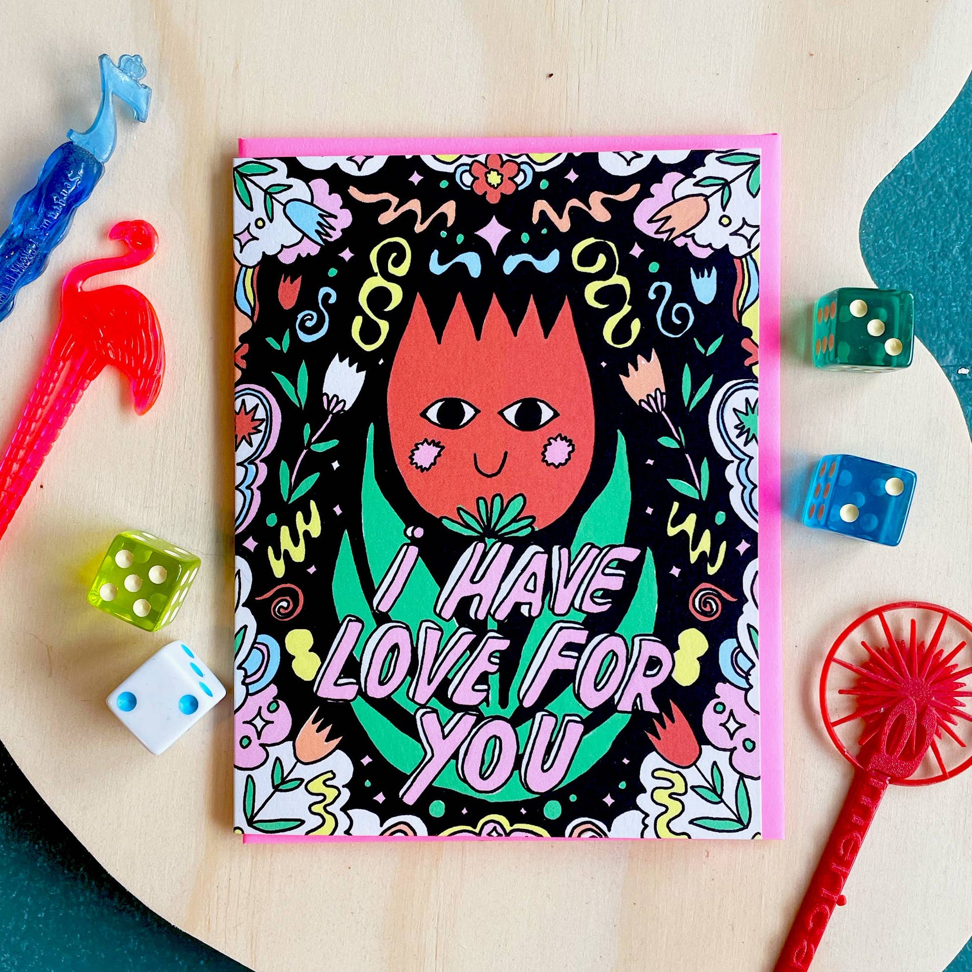 Colorful card with a tulip shaped character and text 'I have love for you' on a wooden surface with toys.
