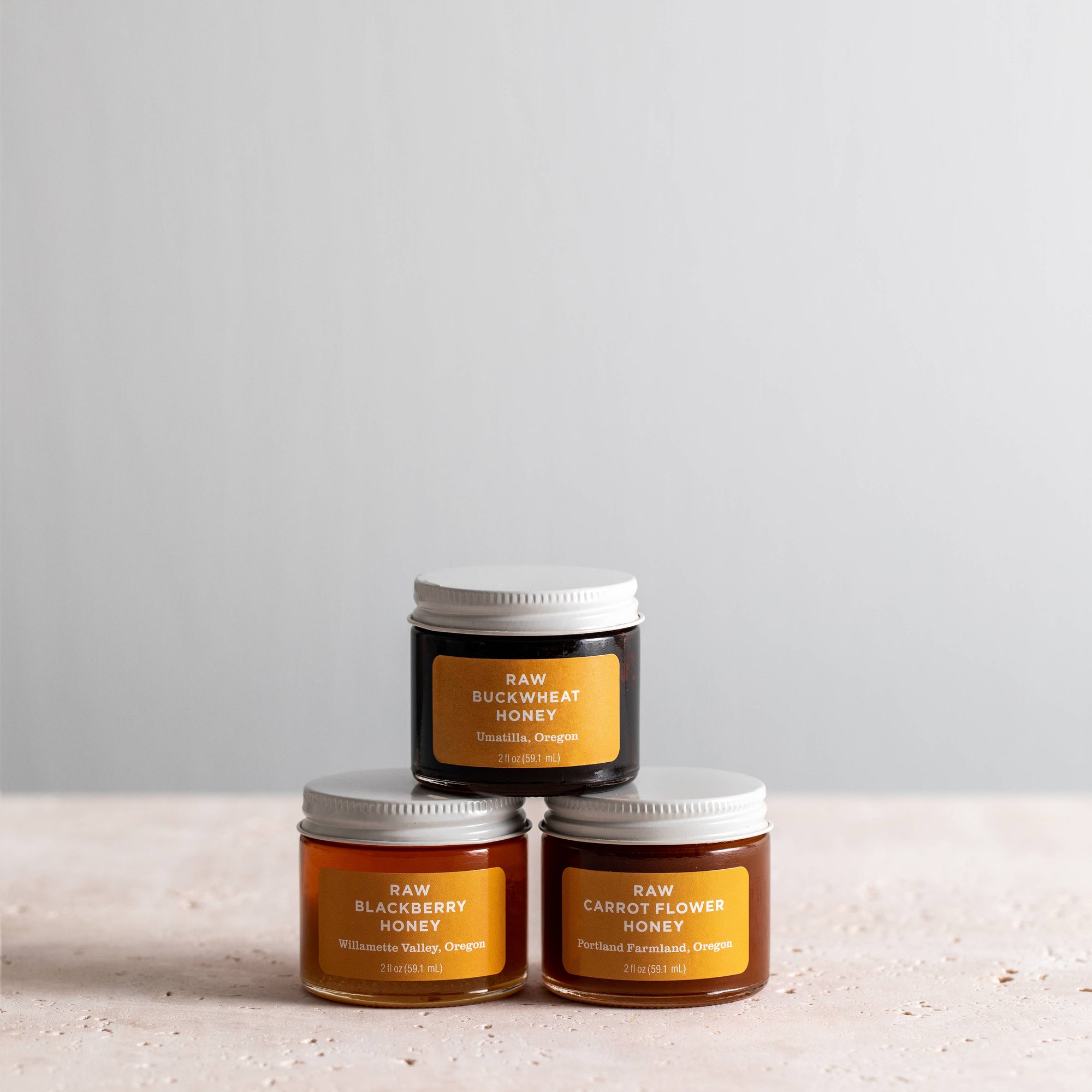 Three small jars of honey arranged in a pyramid on a light stone surface against a soft gray background. The top jar is dark with a yellow label reading RAW BUCKWHEAT HONEY, and the two bottom jars are amber with yellow labels reading RAW BLACKBERRY HONEY and RAW CARROT FLLOWER HONEY.