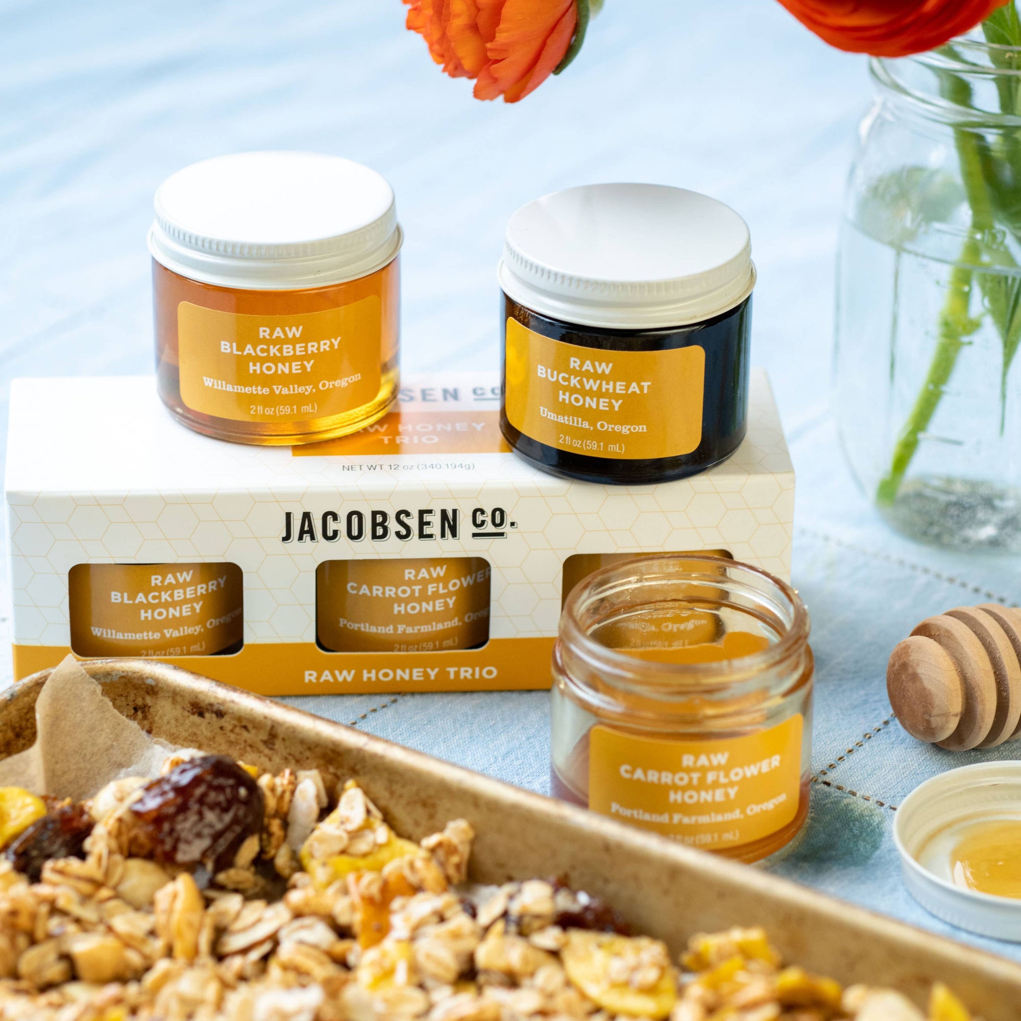 Three glass jars of Jacobsen Co. raw honey displayed on and around a branded box, with a wooden honey dipper and a tray of granola in the foreground, and orange flowers in a glass vase in the background.