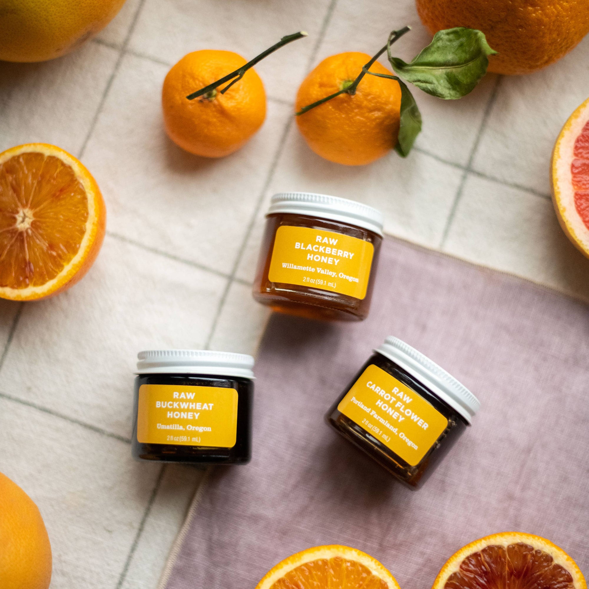 Three small glass jars of honey with yellow labels are arranged on a purple cloth with whole and sliced citrus fruits around them on a light tiled surface.