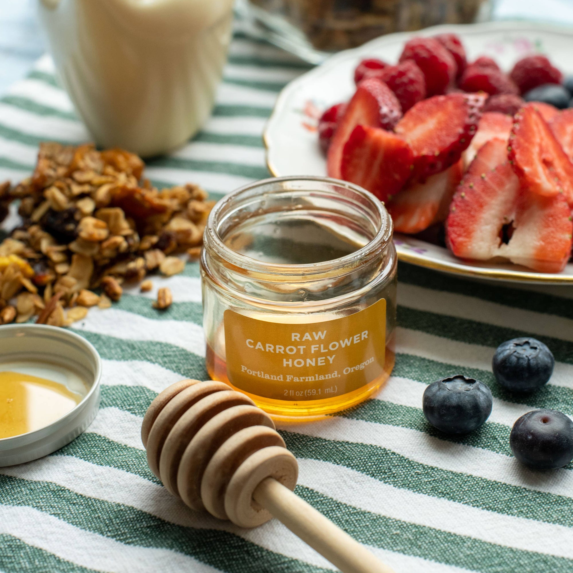 A glass jar of raw carrot flower honey sits on a green and white striped cloth, surrounded by a wooden honey dipper, granola, sliced strawberries, blueberries, and a small cream-colored duck-shaped container.