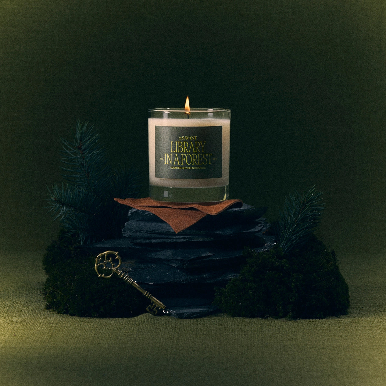 The New Savant Library in a Forest candle with a green label, sitting atop a stack of stones with evergreen branches around it against a dark green background.