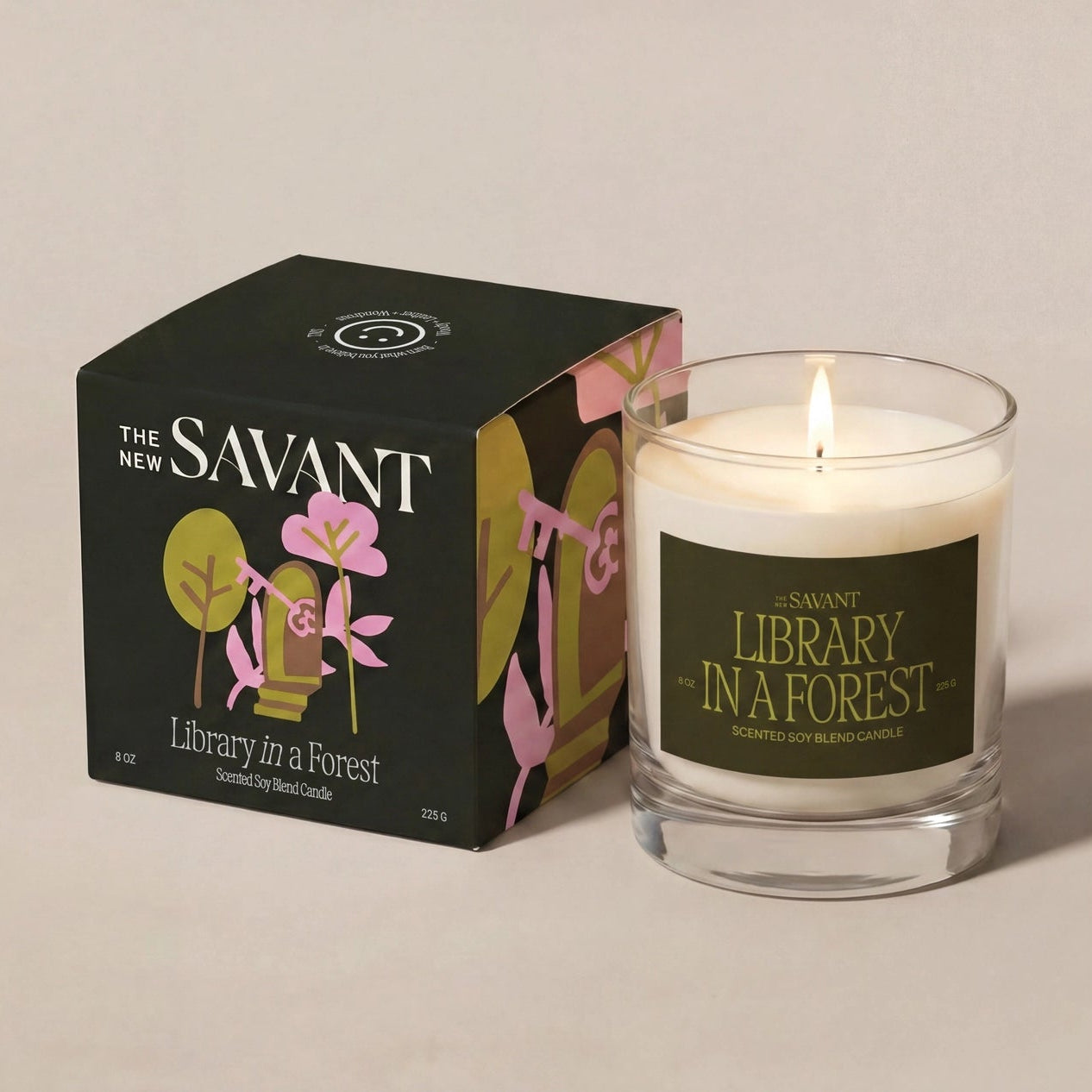 The New Savant 'Library in a Forest' scented candle in a clear glass jar with a lit flame, shown next to its black packaging box with pink and green botanical illustrations on a warm beige background.