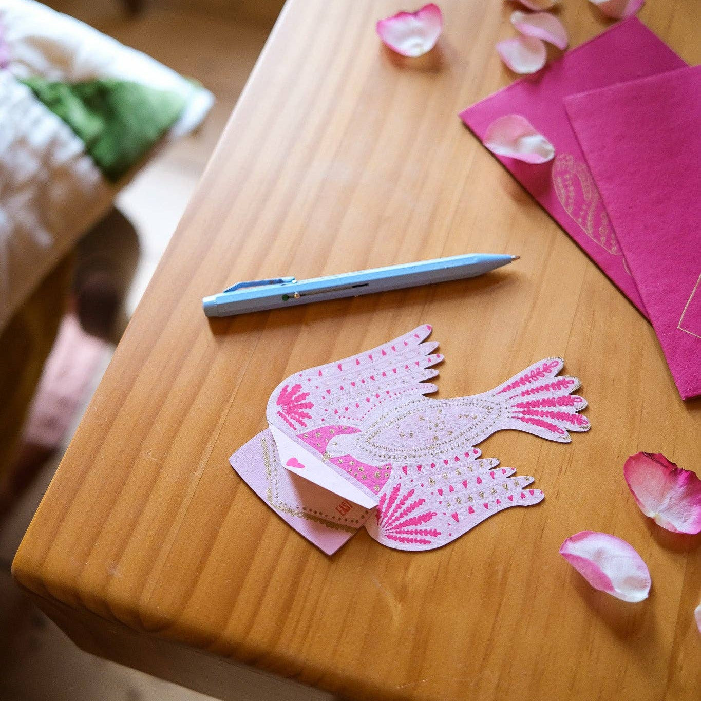 Decorative pink paper bird on a wooden table with rose petals and a pen.