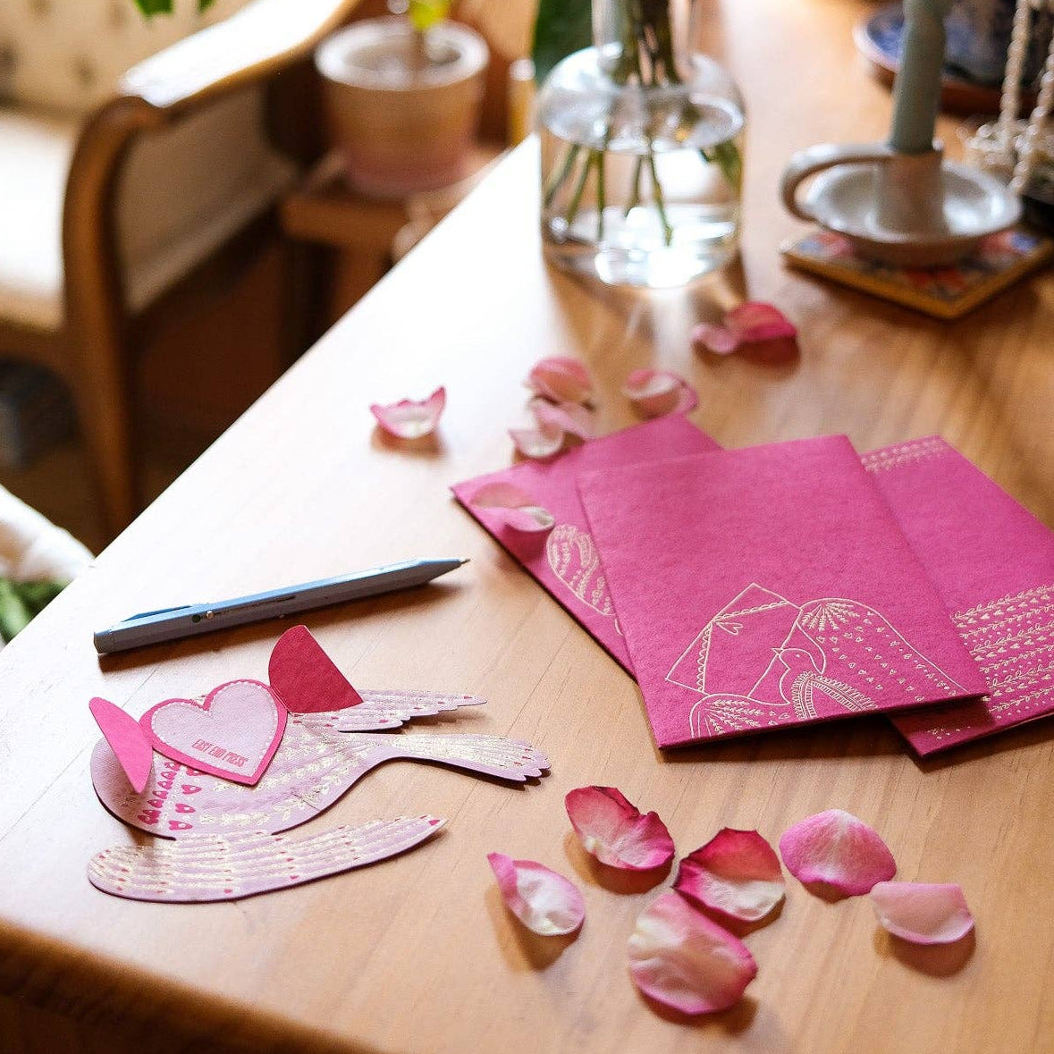 Decorative setup with pink cards, flower petals, and a vase on a wooden table.