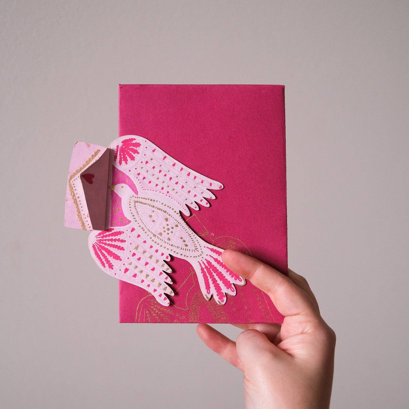 Hand holding a pink card with a decorative bird cutout on a plain background
