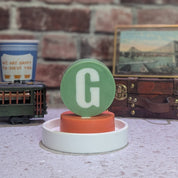 Decorative item with a green letter 'G' on a white base against a brick wall.