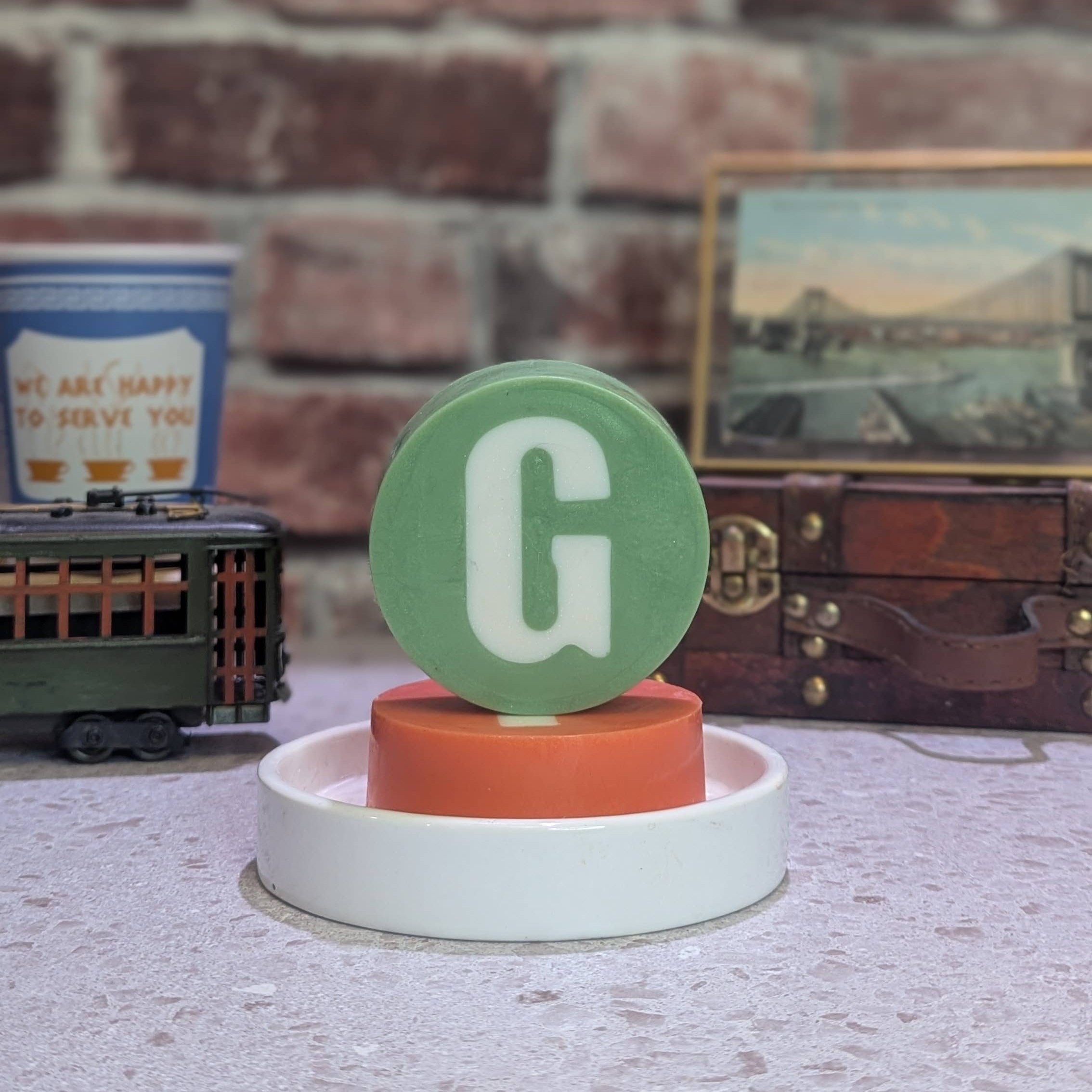 Decorative item with a green letter 'G' on a white base against a brick wall.