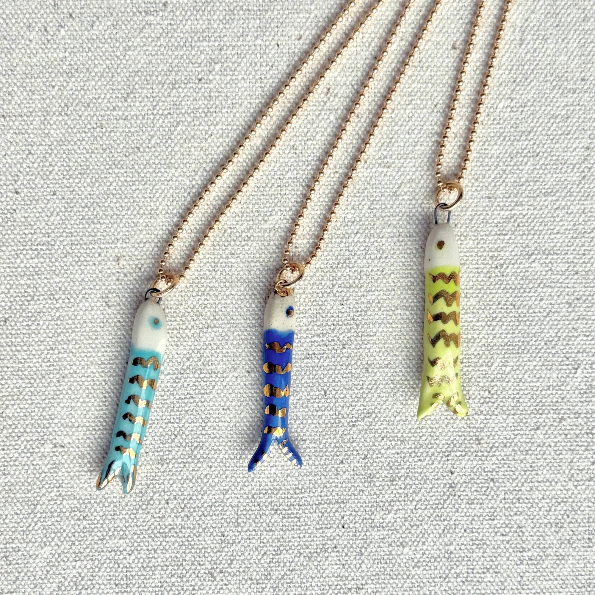 Three fish-shaped pendants on gold chains against a textured gray background