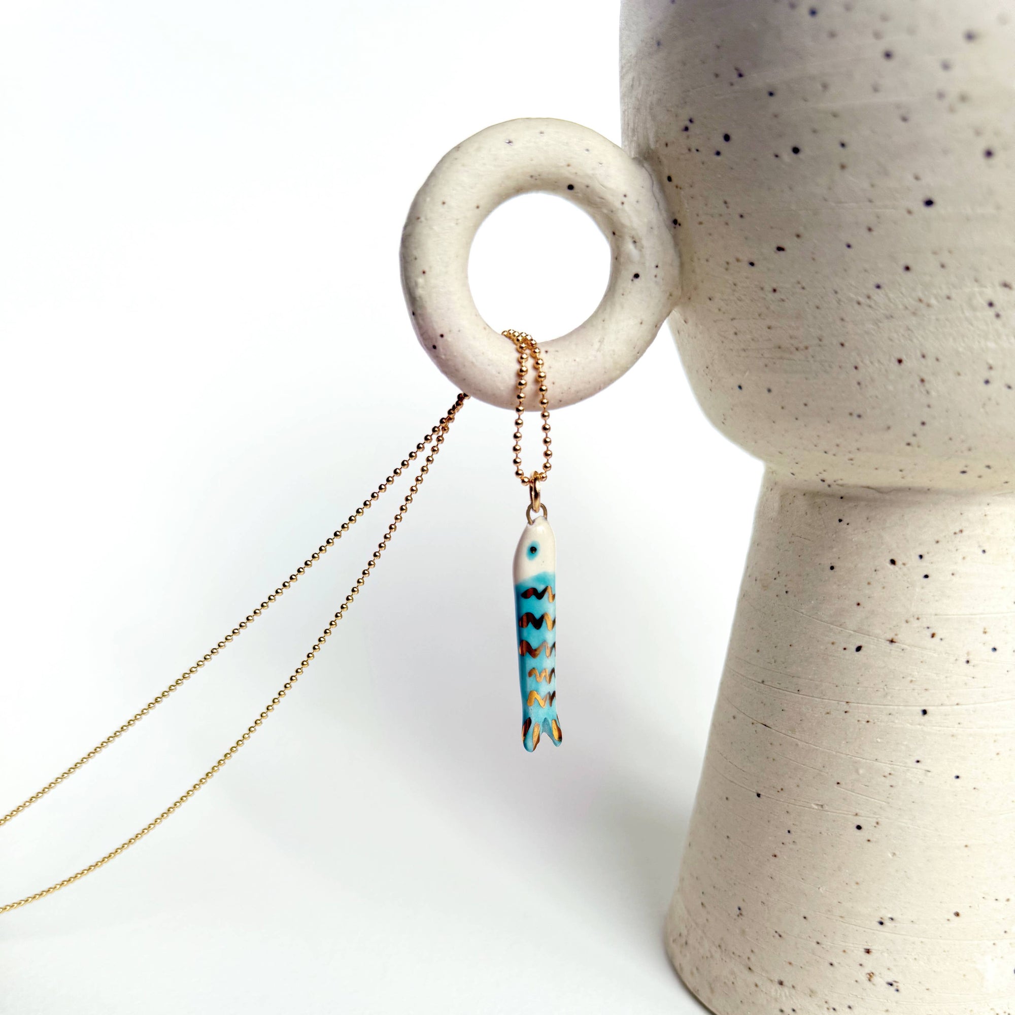 Necklace with a blue fish pendant hanging from a ceramic vase on a white background
