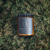 A brown candle with a label that reads 'Moonstone Rituals Forest Witch', placed on a bed of greenery.