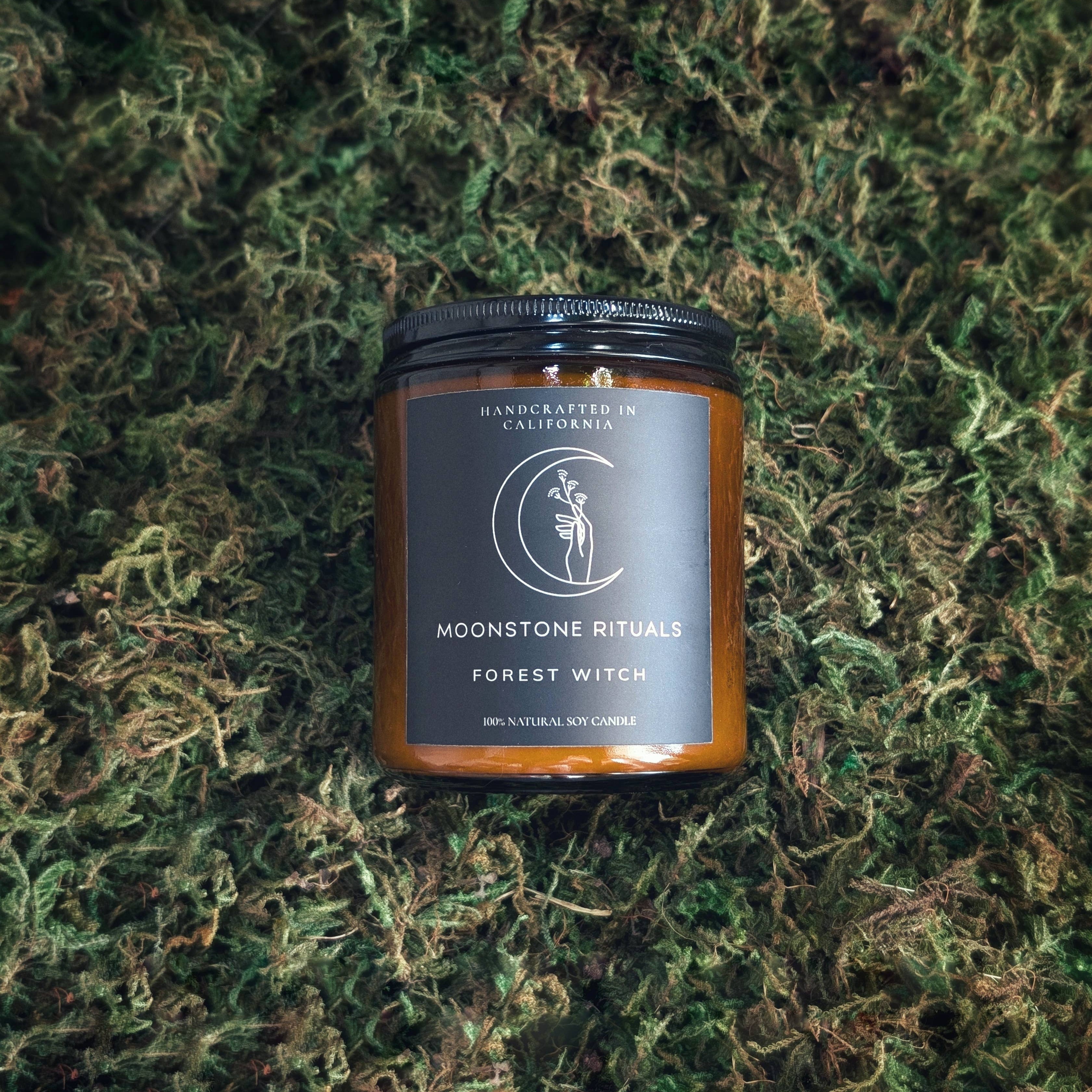 A brown candle with a label that reads 'Moonstone Rituals Forest Witch', placed on a bed of greenery.