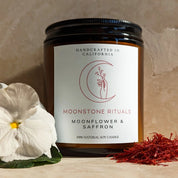 Candle jar with 'Moonstone Rituals' label, white flower, and red herbs on a beige background