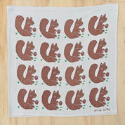 A square white tea towel with a repeating pattern of brown squirrels holding acorns, printed on a light wood background.