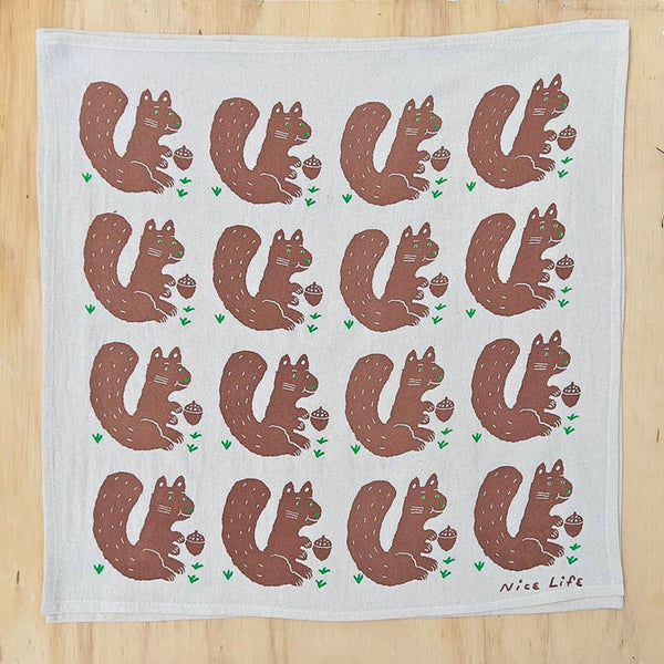 A square white tea towel with a repeating pattern of brown squirrels holding acorns, printed on a light wood background.