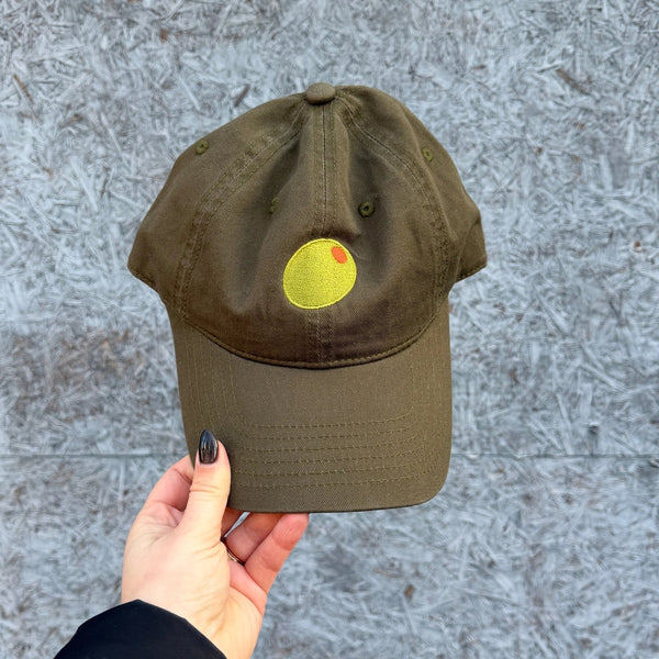 A hand holding an olive green hat with an olive illustration on the front against a gray, concrete background.