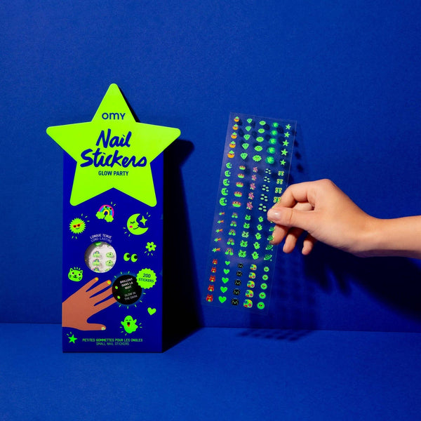 OMY nail stickers packaging with a hand holding a sheet of green glow-in-the-dark stickers against a blue background.