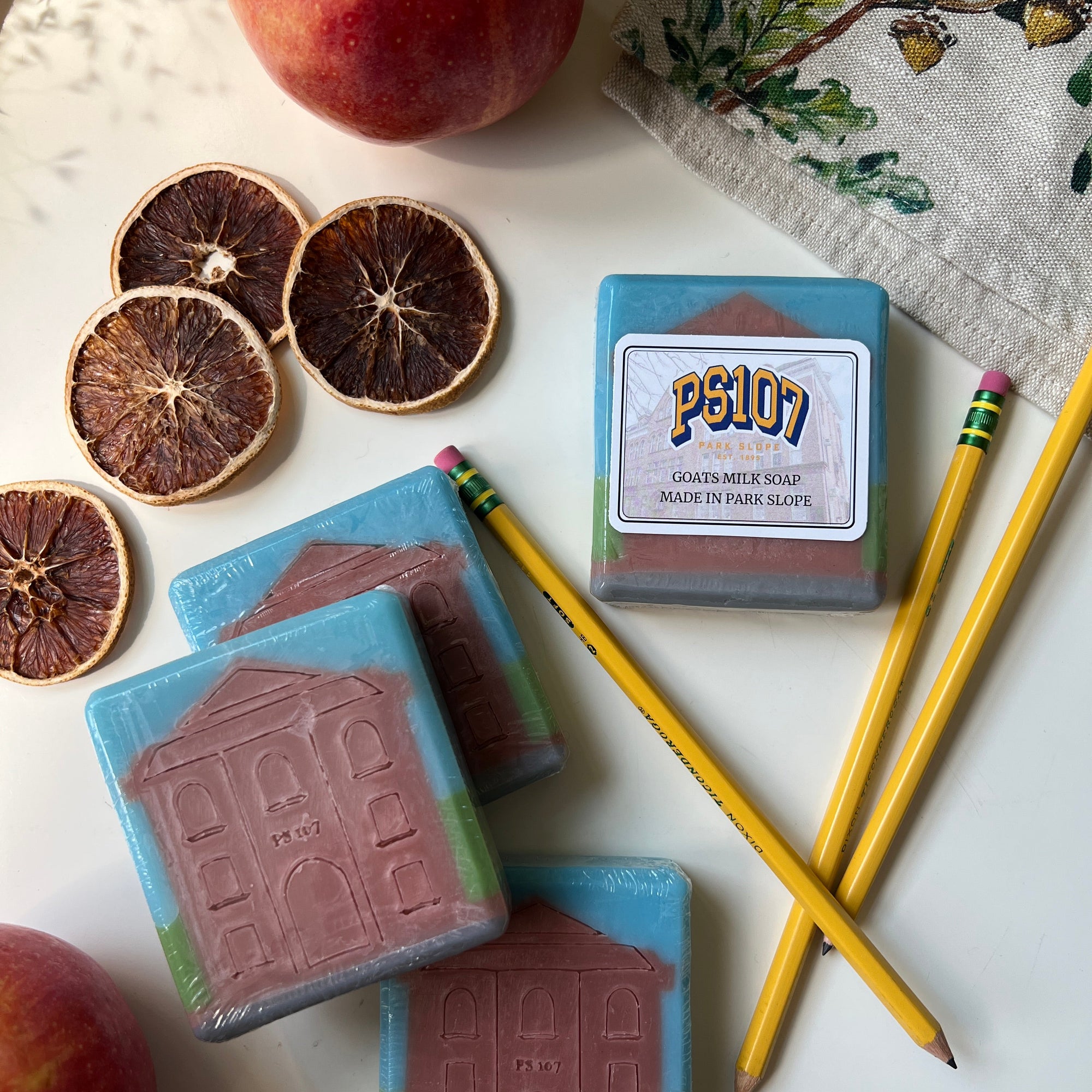 Three bars of soap with a house design, a pencil, an apple, and dried oranges on a white surface.