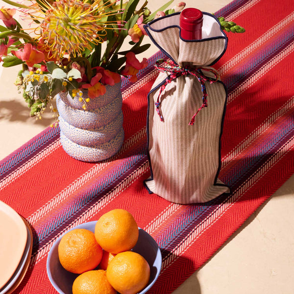 Wine bottle wrapped in cloth with a decorative bow on a red tablecloth with oranges and flowers.