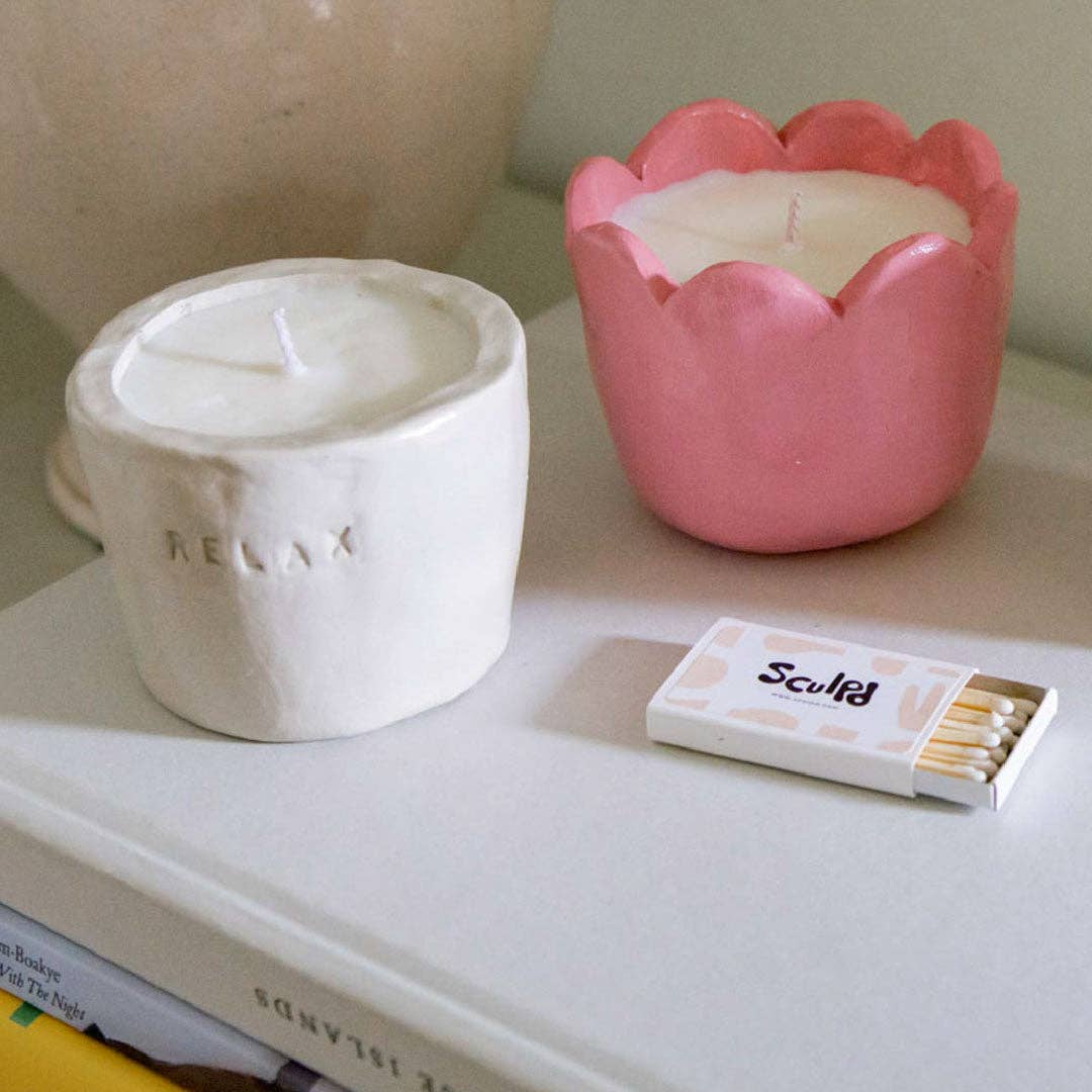 Two candles, one white and one pink, on a surface with a matchbox labeled 'Sculpt'.