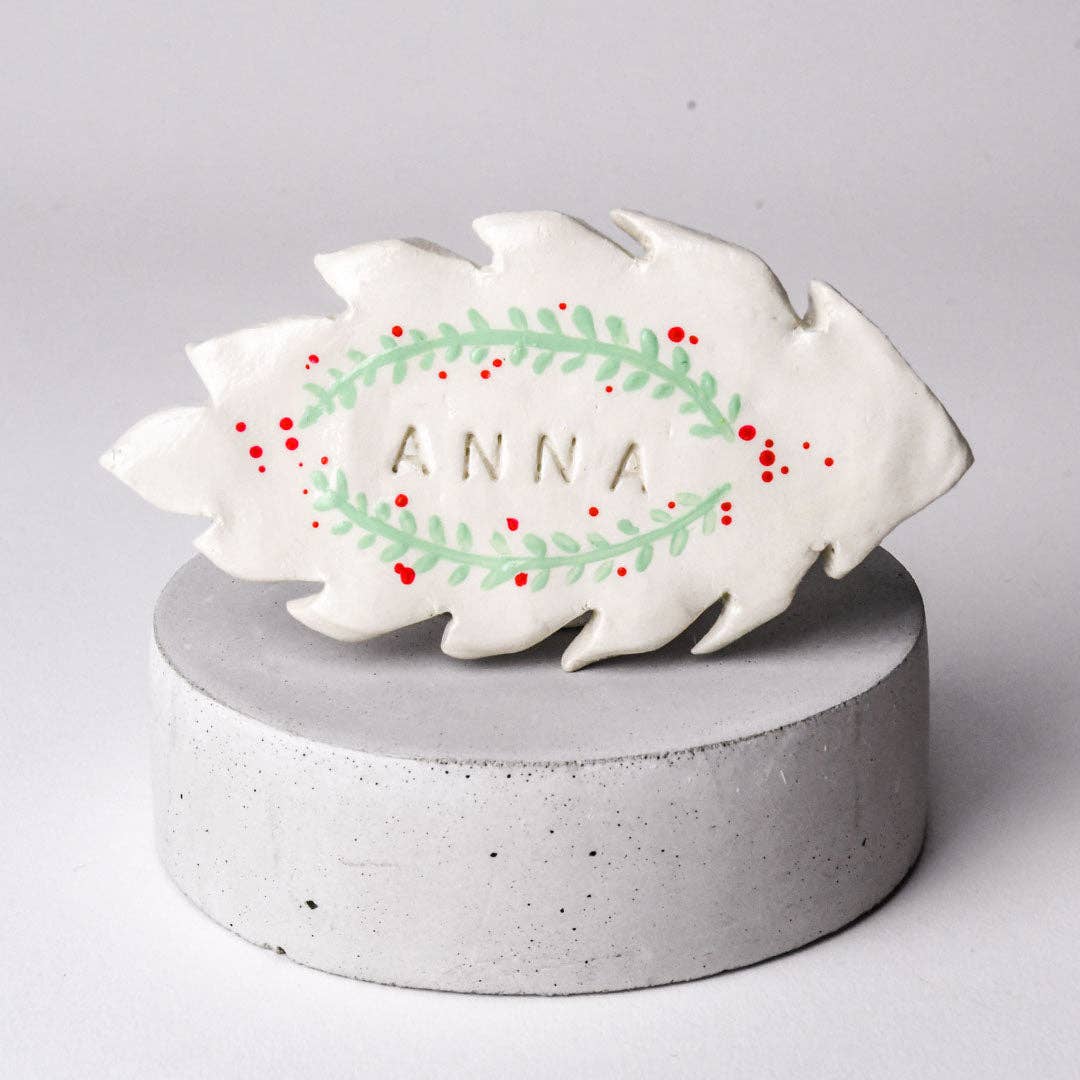 Decorative leaf-shaped ornament with 'ANNA' on a gray pedestal against a light gray background