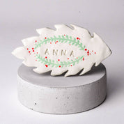 Decorative leaf-shaped ornament with 'ANNA' on a gray pedestal against a light gray background
