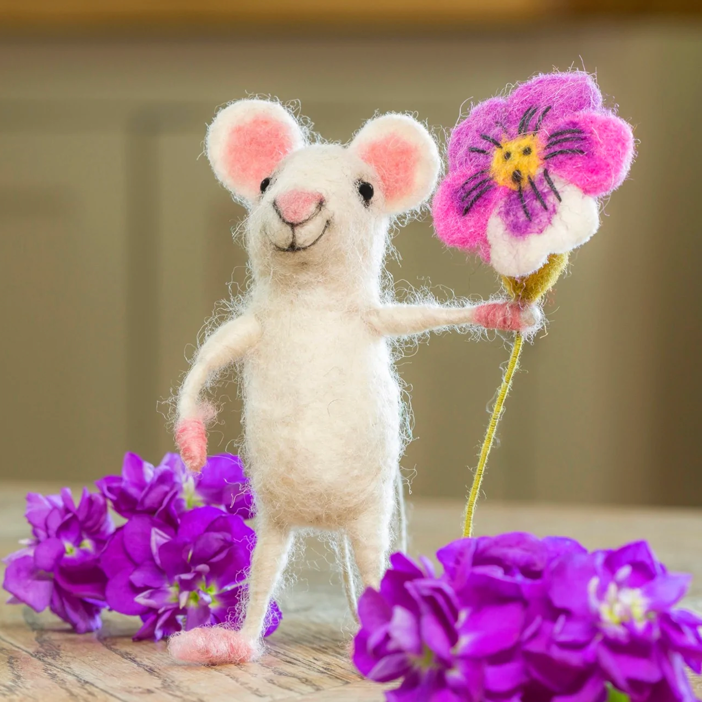 Stuffed felt mouse toy holding a pink flower with purple flowers on a wooden surface.