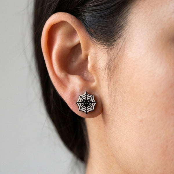 A close-up of a person's ear wearing a spiderweb stud earring with a black spider in the center against a plain light gray background.