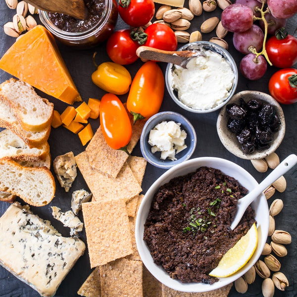 Assorted cheeses, crackers, fruits, and spreads on a slate board with a blue background