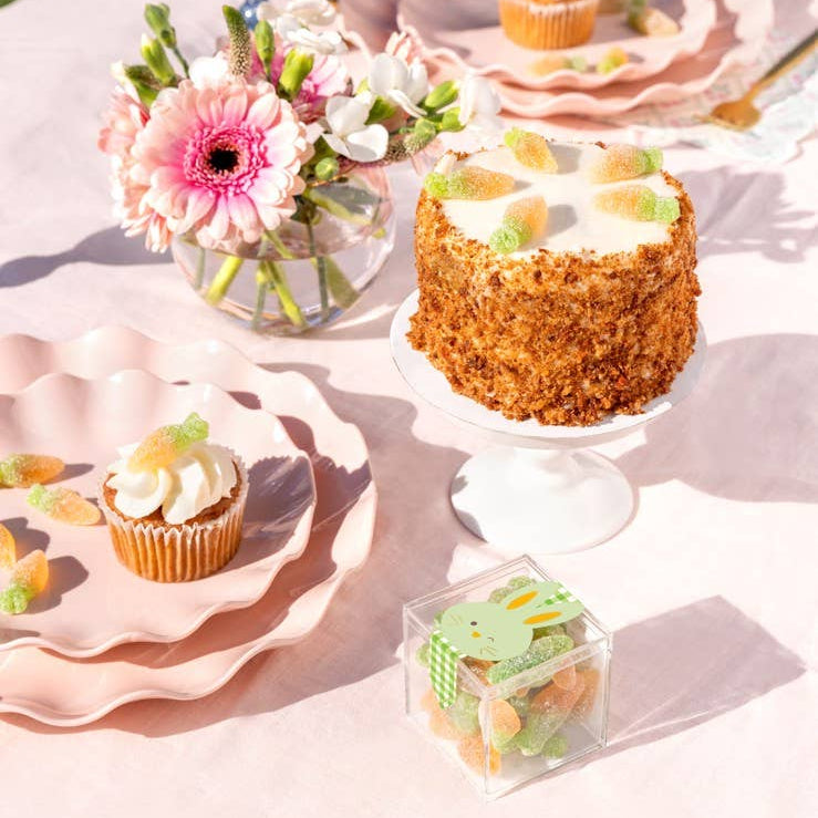 Table setting with a cake, cupcakes, and flowers on a white tablecloth outdoors.