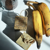 Two bars of soap against a natural background with plantains and a measuring cup with coffee grounds.