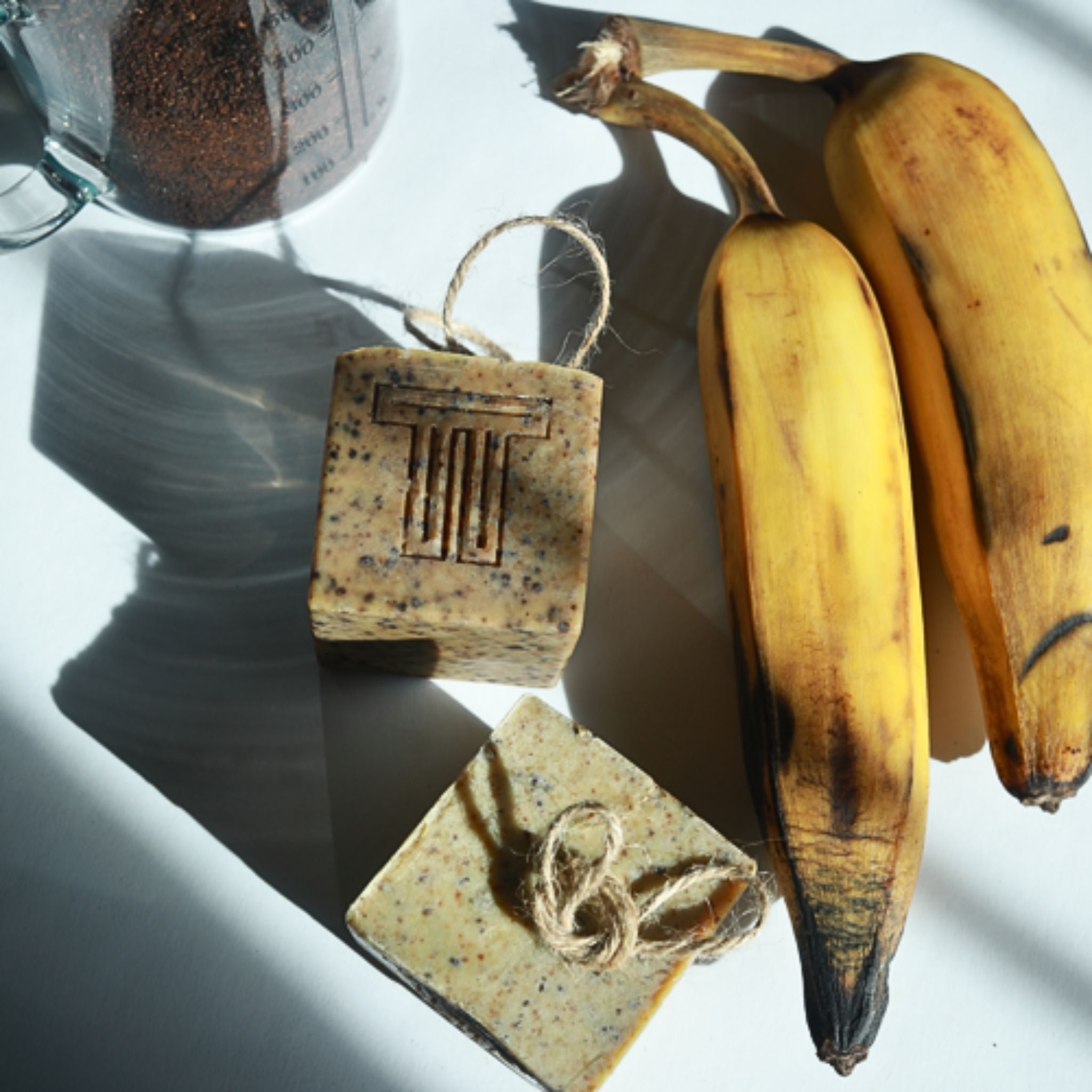 Two bars of soap against a natural background with plantains and a measuring cup with coffee grounds.