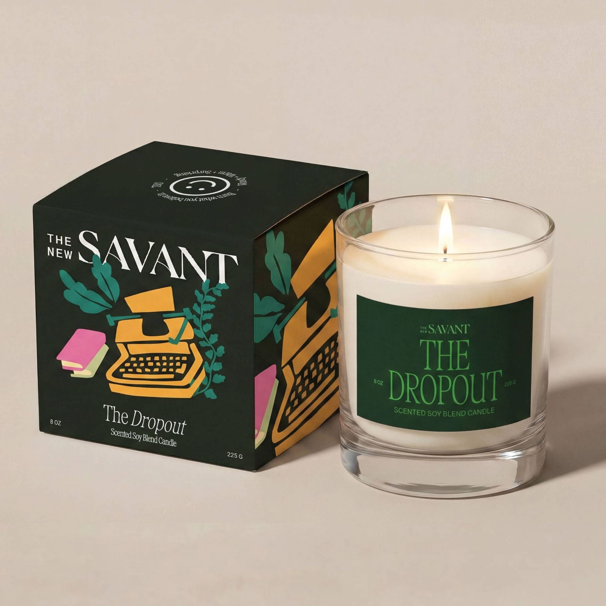 A lit Savant The Dropout scented soy candle in a clear glass jar sits next to its black illustrated box on a warm beige background.