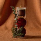 A lit candle in a clear glass jar sits on top of two dark red apples, styled with a eucalyptus sprig and pine branch against a warm brown draped fabric background. The candle label reads 'GIRLINA SWEATER'.