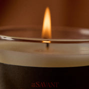 Close-up of a lit candle in a clear glass vessel with a dark label reading 'The New Savant Girl in A' in red text, against a warm brown background.