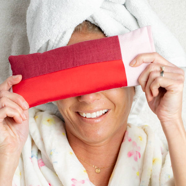 Person wrapped in a towel holding a red and pink eye pillow.
