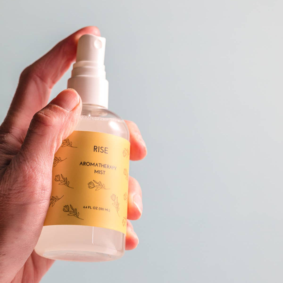 Hand holding a clear spray bottle with a yellow label against a light gray background