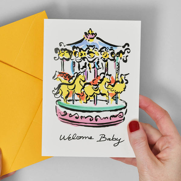 Hand holding a card with a colorful carousel illustration and 'Welcome Baby' text, next to a yellow envelope.