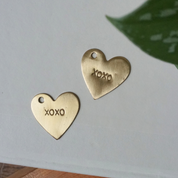 Two gold-colored brass tags in the shape of hearts with 'XOXO' stamped on them, displayed on a white surface with a green leaf in the background.