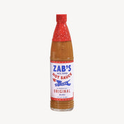 A bottle of Zab's Hot Sauce, Original, featuring a white label with blue and red text, a red cap, and a red patterned neck sleeve, containing a reddish-brown sauce, set against a plain light gray background.