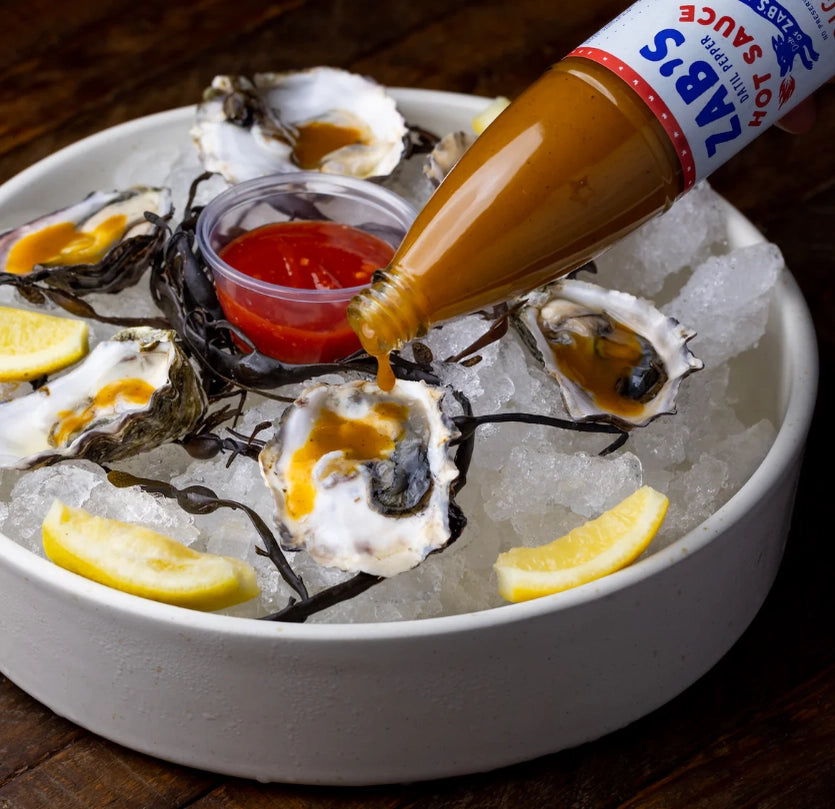 A bottle of hot sauce pouring onto a bowl of oysters served over ice. 